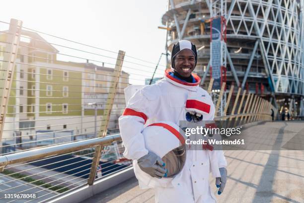 smiling male astronaut holding space helmet while standing on bridge in city - space helmet stock-fotos und bilder