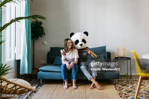 woman wearing panda mask gesturing peace sign while sitting with girlfriend at home - mask disguise stockfoto's en -beelden