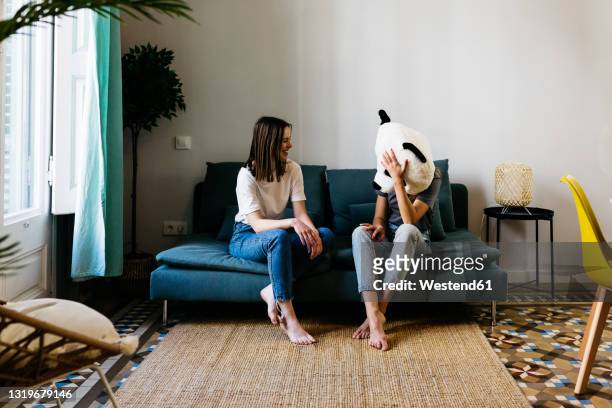 young woman looking at girlfriend wearing panda mask in living room - looking at camera foto e immagini stock
