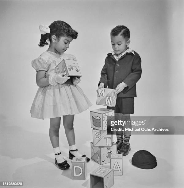 Studio portrait of a young boy and girl, both smartly dressed, as they play with building blocks, against a neutral background, location unspecified,...