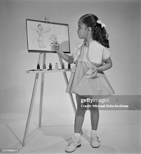 Studio portrait of a young girl drawing on a sheet of paper on an easel, against a neutral background, location unspecified, April 1951. Image shot...