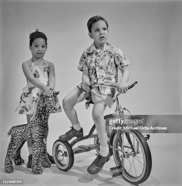 Studio portrait of a young girl with a stuffed toy alongside a young boy sitting on a tricycle, against a neutral background, location unspecified,...