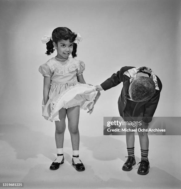 Studio portrait of a young boy lifting the hem to look under a young girl's dress, both children are smartly dressed, against a neutral background,...