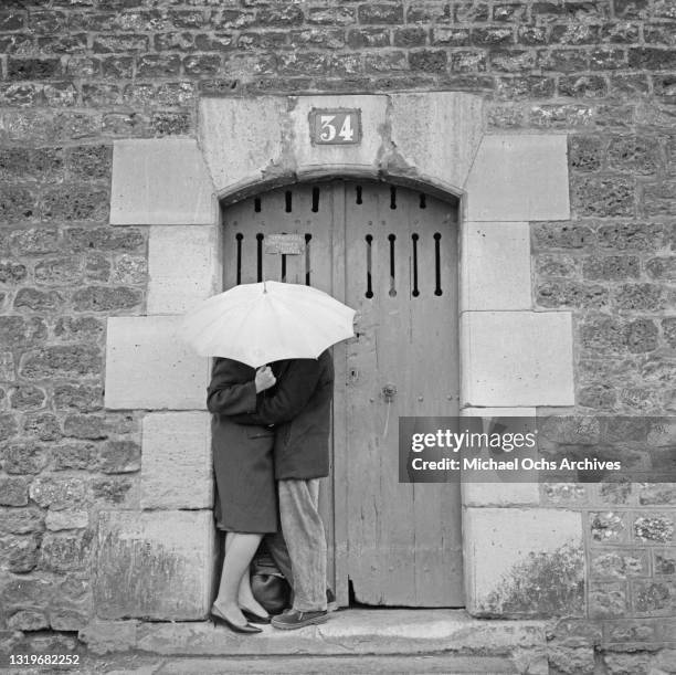 Kissing couple sheltered from the rain by an umbrella, standing by a doorway with the number 34 on an unspecified street in Paris, France, March 1961.