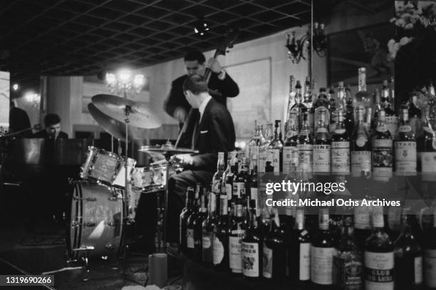 An unspecified piano trio, with the drummer obscuring the bassist, performing beside the bar at an unspecified nightclub in New York City, New York,...