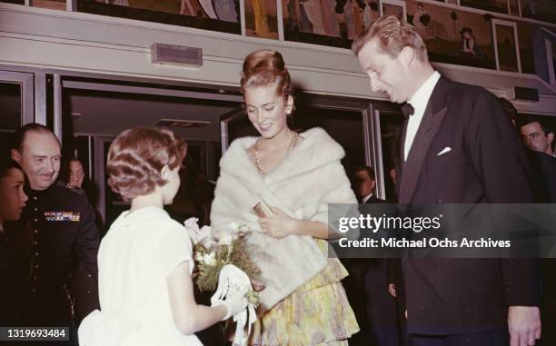 Young girl presents a bouquet of flowers to Belgian Royal Paola, Princess of Liege, wearing a yellow, green and pink outfit with a gold necklace and...