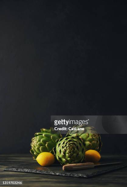 stilleven achtergrond met drie artisjokken, citroen en keukenmes op een lei snijplank - clair obscur stockfoto's en -beelden