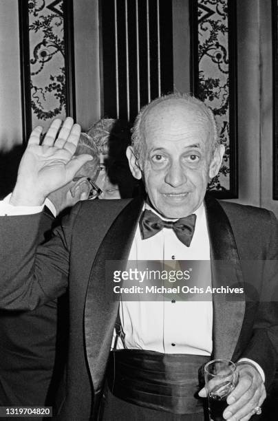 An unspecified elderly man, wearing a tuxedo with a bow tie and cummerbund, raising his right hand as if waving, with a drink in his left hand, at...