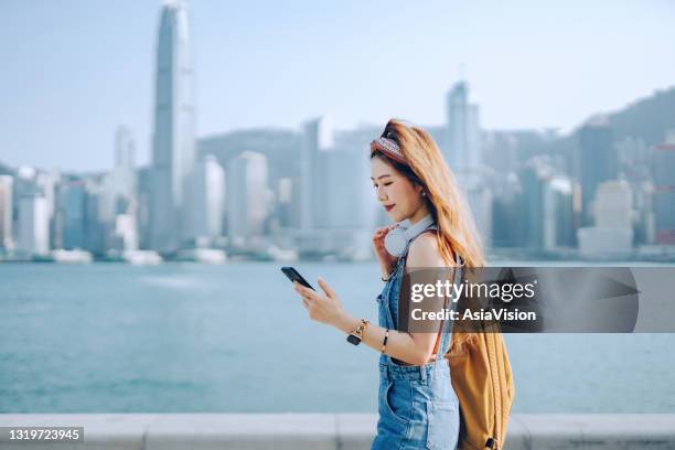 schöne junge asiatische frau trägt rucksack und trägt kopfhörer um den hals. sie ist sms auf dem smartphone, während sie entlang der promenade gegen spektakuläre stadt skyline an einem sonnigen tag. lifestyle und technologie - hong kong girl stock-fotos und bilder