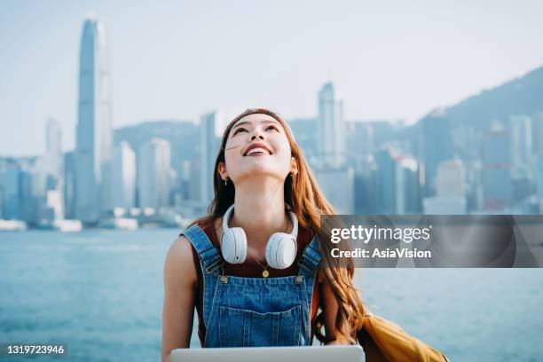 schöne lächelnde junge asiatische frau sitzt an der promenade des victoria hafen, gegen urbane stadt skyline. sie trägt kopfhörer um den hals, mit laptop. mit hoffnung und positiver emotion in den himmel schauen. lifestyle und technologie - hong kong girl stock-fotos und bilder