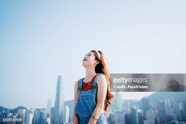 美しい笑顔の若いアジアの女性は、ビクトリア港の遊歩道で壮大な都市のスカイラインに対して、屋外で新鮮な空気と穏やかな風を楽しみながら、深呼吸しながら彼女の目を閉じました - very young asian girls ストックフォトと画像