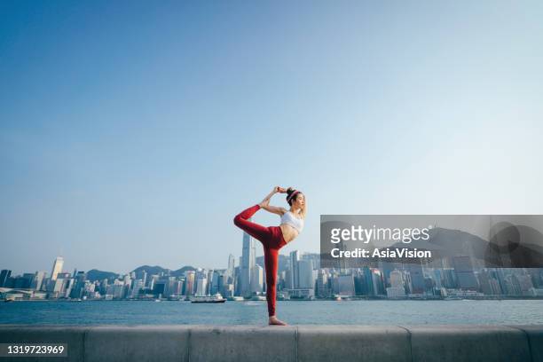 an active young asian sports woman practicing mindfulness yoga outdoors in the morning, against spectacular hong kong city skyline by the promenade of victoria harbour. girl power, welling, fitness and healthy lifestyle concept - commercial buildings hong kong morning stock pictures, royalty-free photos & images