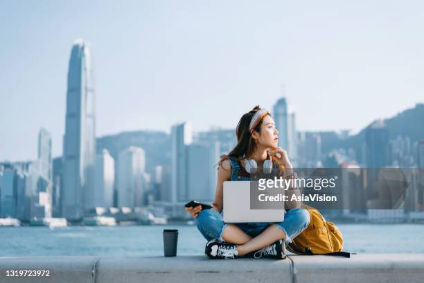 美麗的亞洲年輕女子盤腿坐在長廊旁，與城市天際線對立。她戴著耳機，使用智能手機，在筆記型電腦上工作，身邊有一個咖啡杯。在思想中望向遠方。生活方式和技術 - hong kong 個照片及圖片檔