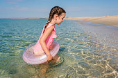 Happy cute tollder girl 5-6 years old in bright pink swimsuit with swim tubes ring going into the sea for swimming on sunny day. Kids having fun at the beach. Family vacation with children concept