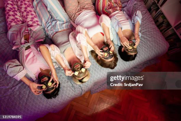 hoge hoekmening van vier jonge vrouwen die op het bed met gezichtsschoonheidsmaskers en plakken komkommers op hun liggen - party mask stockfoto's en -beelden