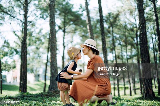 side view of a loving young asian mother kissing and embracing her lovely little daughter in the woods, against beautiful sunlight. precious moment between mother and daughter. family lifestyle. family love and care. exploring in nature and outdoor fun - asian mom kid kiss stock-fotos und bilder