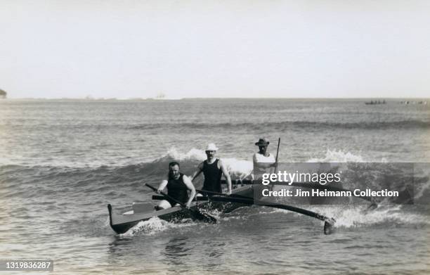 Black-and-white photo shows three men in an outrigger canoe riding a wave towards the beach at Waikiki, circa 1921.