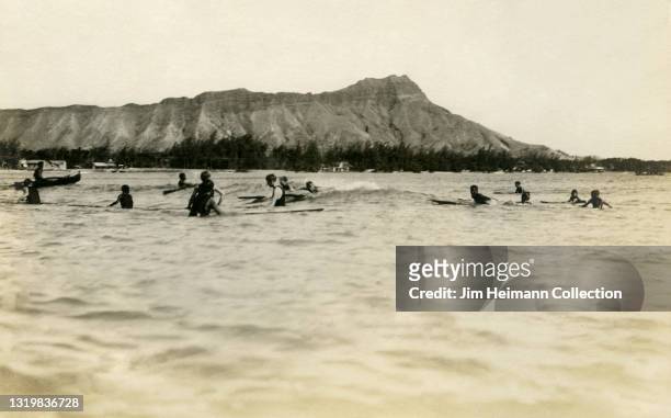 Black-and-white film photo shows a group of surfers paddling out at Waikiki with Diamond Head in the background, circa 1916.