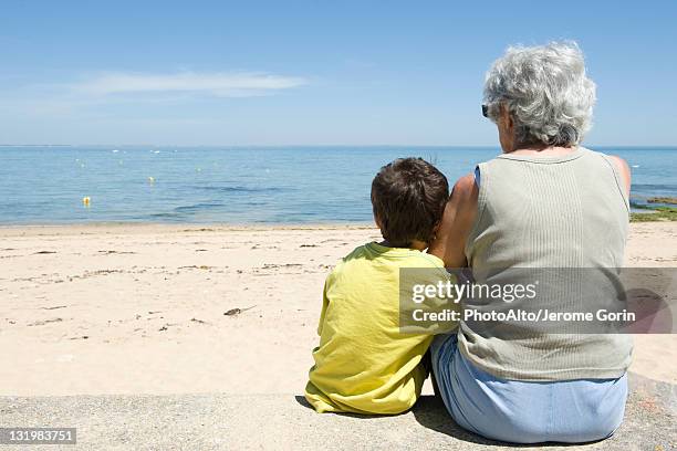 grandmother and grandson sitting together on beach, looking at sea - grandson stock pictures, royalty-free photos & images