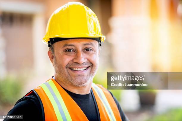 lächelnder hispanischer bauarbeiter mit arbeitshelm, der in die kamera schaut - hispanic construction worker stock-fotos und bilder