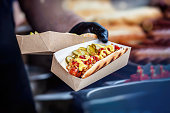 Hand of chef holding cardboard box with freshly prepared hot hot dog with tasty additions. Barbecue, grill concept
