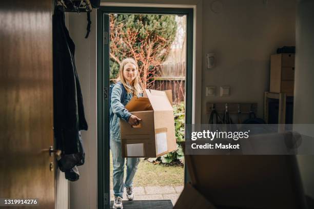 full length of young woman carrying box while walking in through doorway - entrando fotografías e imágenes de stock