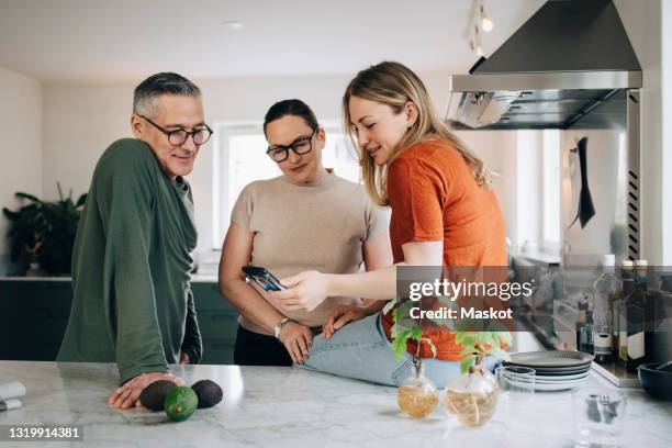 young woman discussing over smart phone with parents while sitting on kitchen island at home - mother daughter kitchen stockfoto's en -beelden