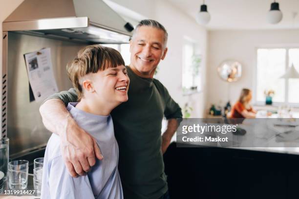 Mature man with arm around looking at cheerful son in kitchen