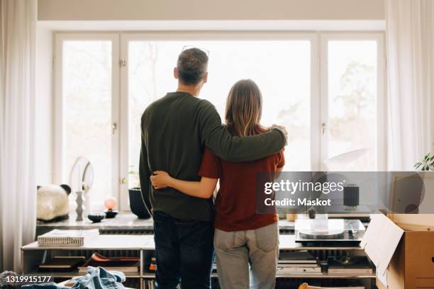 rear view of father and daughter standing with arms around while looking through window at home - woman window home stock pictures, royalty-free photos & images