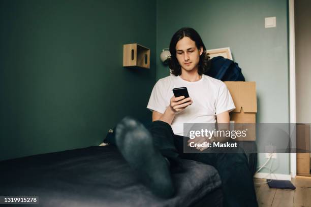 young man using smart phone while sitting on bed at home - alleen één man stockfoto's en -beelden