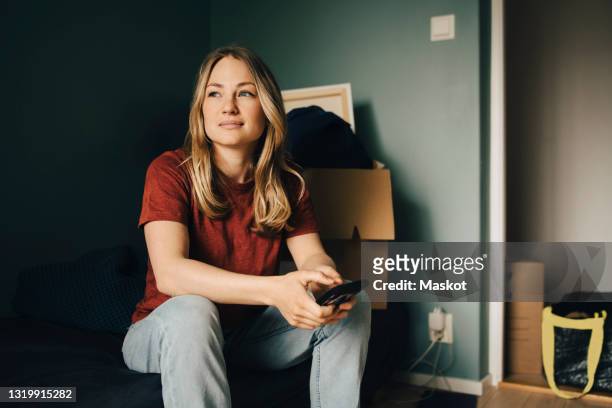 thoughtful young woman looking away while sitting with smart phone in bedroom - beschouwing stockfoto's en -beelden