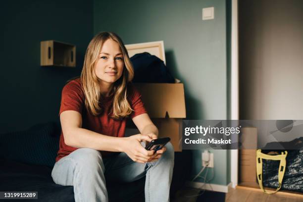 portrait of young woman sitting with smart phone in bedroom at home - portrait mobilephone stock pictures, royalty-free photos & images
