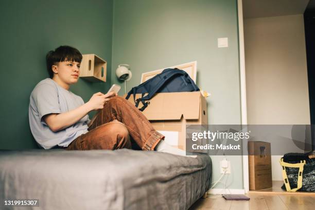 thoughtful transgender person sitting with smart phone on bed while looking away in bedroom - boomerang generation stock pictures, royalty-free photos & images