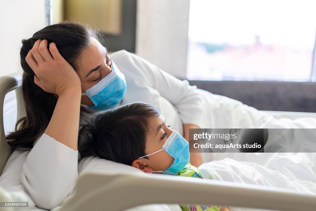 Tired sleeping mother lying on the hospital bed with sick kid wearing protective face mask