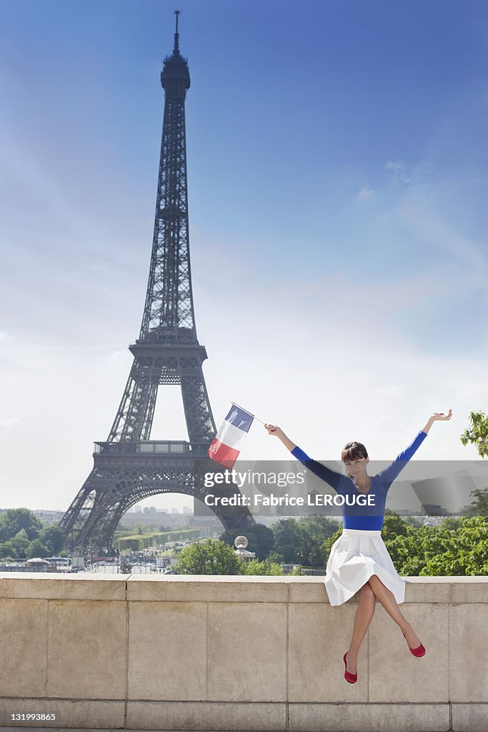 Woman holding a French flag sitting on a stone wall with the Eiffel Tower in the background, Paris, Ile-de-France, France