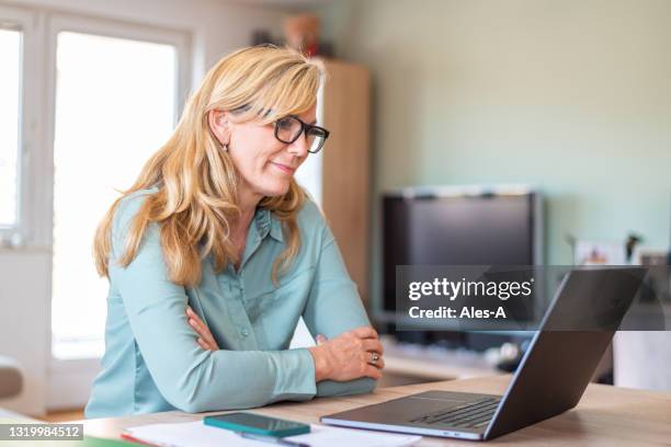 mature woman using laptop while sitting at the table at home - unhappy woman blonde glasses stock pictures, royalty-free photos & images