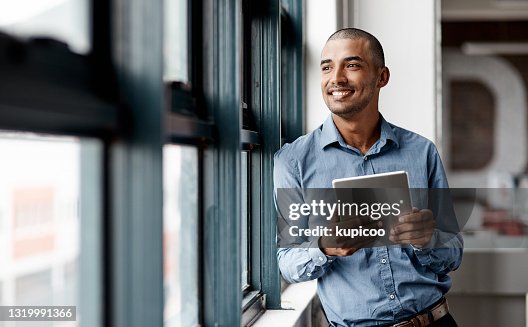 Shot of a young businessman using a digital tablet while standing at a window in an office Shot of a young businessman using a digital tablet while standing at a window in an office