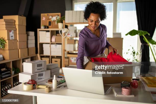 imprenditrice che usa il laptop mentre impacchetta i vestiti in una scatola di cartone - e commerce foto e immagini stock