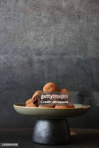 image of homemade, individual chocolate truffle sweets with ganache centre piled on cake stand with bite taken out of one, luxury chocolate balls coated in dusting of cocoa powder, dark grey background, focus on foreground - milk chocolate truffles stock pictures, royalty-free photos & images