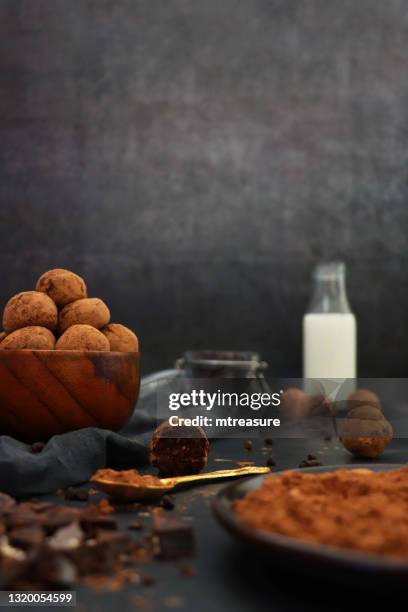 image of homemade, individual chocolate truffle sweets with ganache centre, pile of luxury chocolate balls coated in dusting of cocoa powder in a wooden bowl beside grey tea towel, metal spoon, milk bottle, dark grey background, focus on foreground - milk chocolate truffles stock pictures, royalty-free photos & images