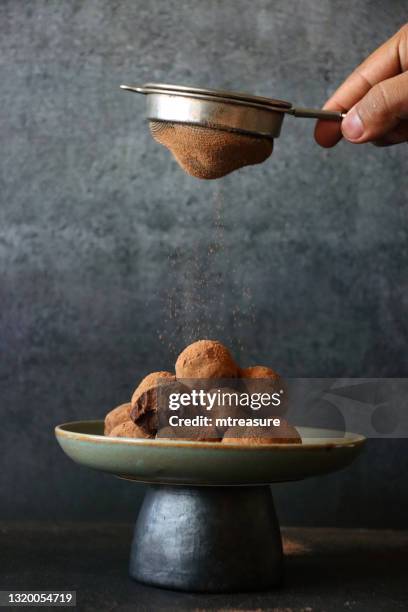 image des sucreries individuelles faites maison de truffe de chocolat avec le centre de ganache empilés sur le stand de gâteau avec la morsure prise d’un, personne méconnaissable passant au crible la poudre de cacao par un tamis en métal saupoudrant  - sifting stock photos et images de collection