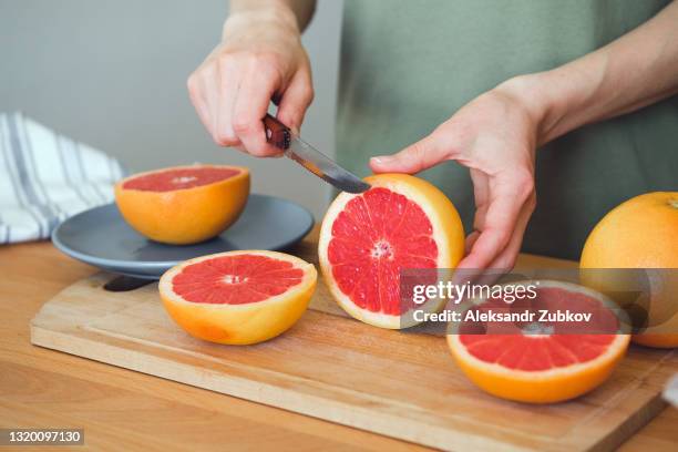 a girl or woman is holding a knife, cutting a red ripe grapefruit in half, on a cutting board, against the background of a wooden kitchen table. there is a manual juicer nearby. making juice for breakfast. the concept of vegetarian, vegan and raw food. - grapefruit red - fotografias e filmes do acervo