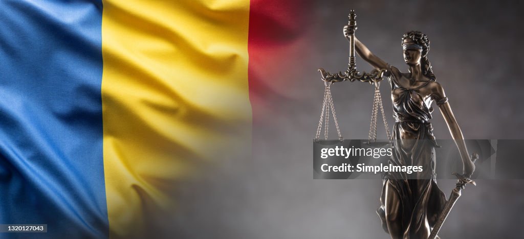 Justice is blind statue symbolizing law with scales and sword in hands and a Romanian flag in the background.