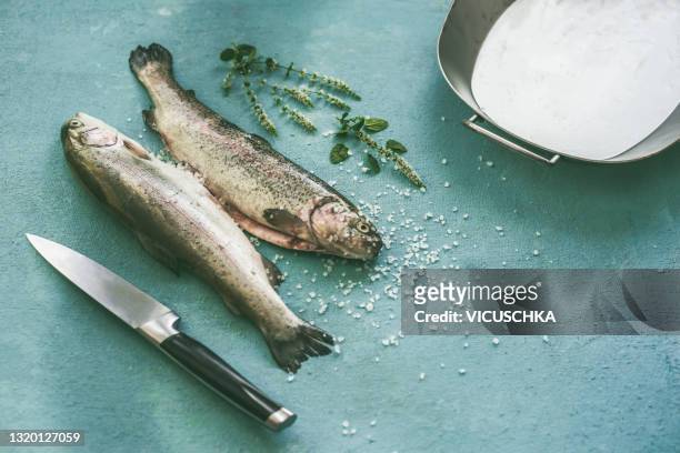 close up of whole trouts with salt, herbs, kitchen knife and baking dish on pale blue background - trout stock pictures, royalty-free photos & images