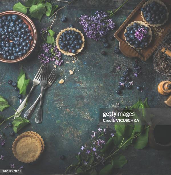 food background frame with blueberry tarts, lilac flowers, rustic forks and bowl with blueberrys - food photography dark background blue stock-fotos und bilder