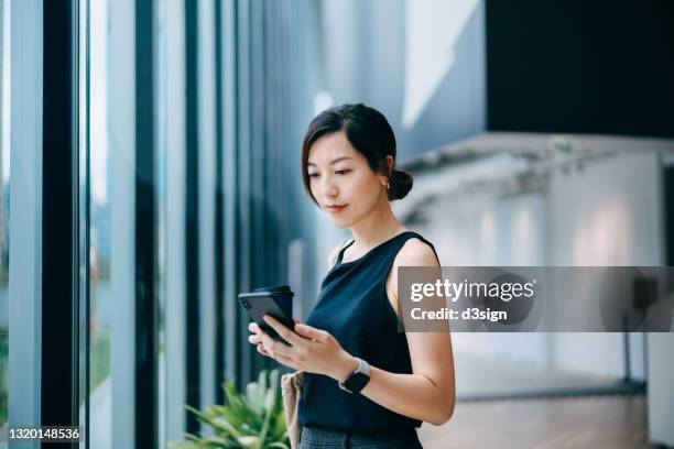 confident and modern young asian businesswoman with coffee to go, standing by window, checking emails on smartphone in a contemporary office space. she is wearing smart casuals in office. female leadership. business on the go - asian and indian ethnicities smartwatch phone stock pictures, royalty-free photos & images