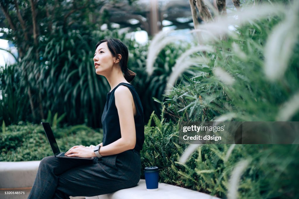 Young Asian businesswoman working on laptop outdoors, sitting on the bench in an urban office park, surrounded by green plants. Looking away in thought. Outdoors with technology. Remote working concept