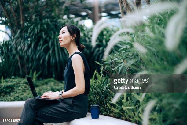 young asian businesswoman working on laptop outdoors, sitting on the bench in an urban office park, surrounded by green plants. looking away in thought. outdoors with technology. remote working concept - optimistic inspiring movement stock pictures, royalty-free photos & images