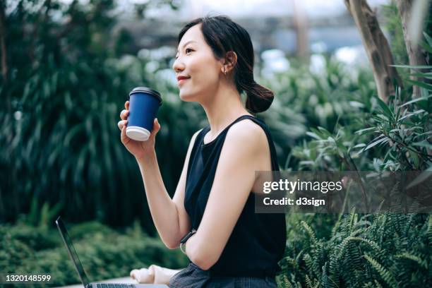 young asian businesswoman looking away while sitting on the bench in urban office park, surrounded by green plants. drinking a cup of coffee while working on laptop in the middle of a work day. outdoors with technology. remote working concept - lunch break stockfoto's en -beelden