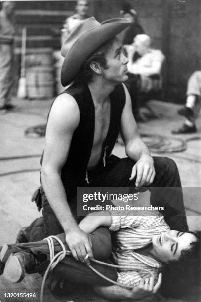 View of American actor James Dean , a cigarette in his hand, and English-American actress Elizabeth Taylor , tangled in a lasso on the floor, during...